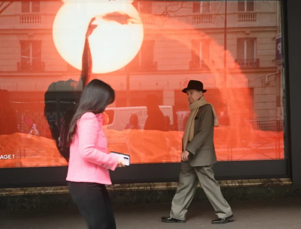 Mary P · 07/03/2026 · 2784 x 2113 Cette photographie montre deux personnes en mouvement devant une vitrine. La femme, en premier plan, porte une veste rose et tient un appareil photo, tandis que l'homme, en arrière-plan, arbore un chapeau et une veste beige.La composition joue sur les contrastes entre les sujets et l'image reflétée, une scène orange et sombre. Le cadrage est serré sur les personnages, avec une lumière naturelle douce. Les couleurs dominantes sont les tons chauds (orange, rose) et neutres (beige, gris).Le style est réaliste et spontané, créant une ambiance urbaine et dynamique.