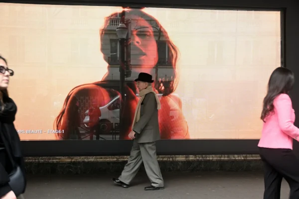 Mary P · 07/03/2026 · 2784 x 1856 Cette photographie montre deux personnes marchant devant une grande affiche publicitaire. Le sujet principal est l’affiche représentant un visage masculin stylisé, aux traits floutés et aux couleurs vives, notamment des tons rouges et oranges.La composition est dynamique, avec un cadrage serré sur les passants et l’affiche en arrière-plan. La lumière semble naturelle, bien que contrastée, mettant en valeur les contours des sujets. Les couleurs dominantes sont chaudes, avec des rouges et oranges sur l’affiche, contrastant avec les tons neutres des vêtements des passants. L’ambiance est urbaine et moderne, évoquant une ville animée.