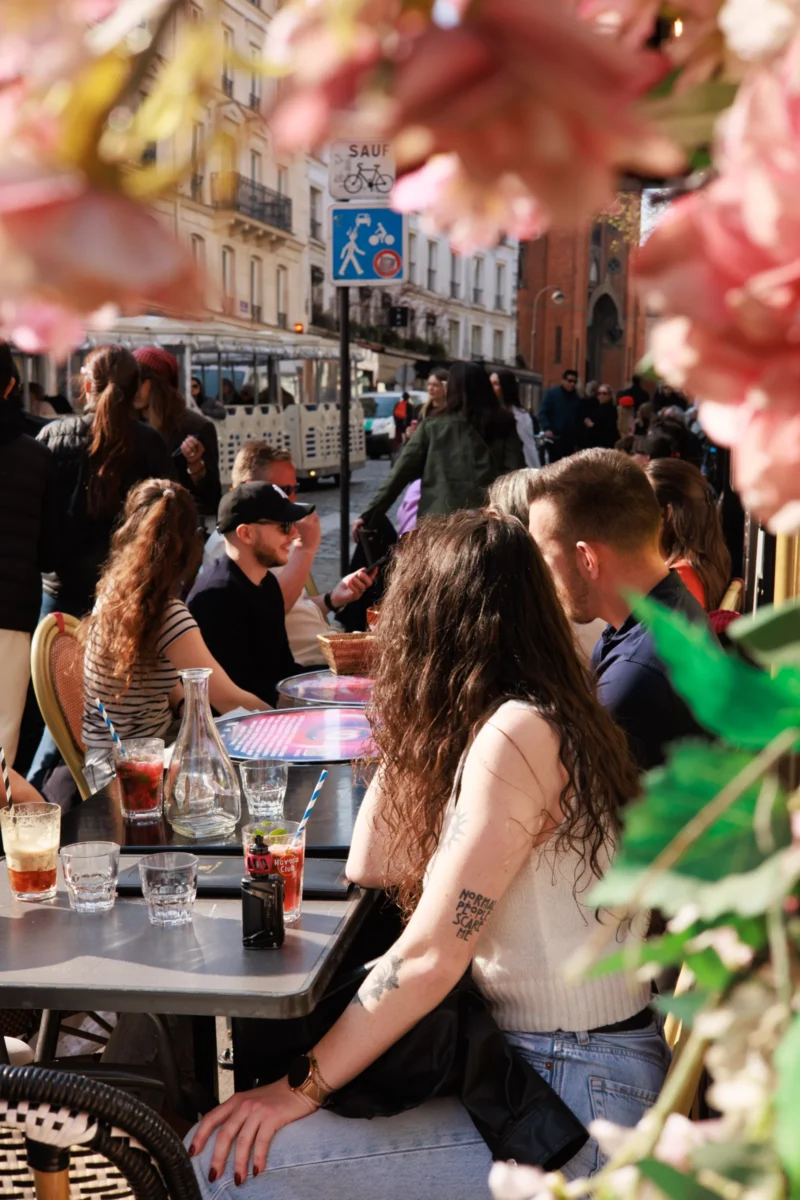 Claude · 3930 x 5895 Cette photographie capture un groupe de personnes en extérieur, assis autour de tables dans un café animé. Le sujet principal semble être une femme avec des cheveux longs, tournée vers son compagnon.La composition est dynamique, avec un cadrage serré sur les personnes en premier plan et un arrière-plan flou mais visible. La lumière est naturelle et douce, typique d’une journée ensoleillée. Les couleurs dominantes sont des tons chauds et naturels : verts, bruns, et teintes pastel des vêtements.Le style est documentaire et spontané, évoquant une ambiance conviviale et détendue, presque estivale.