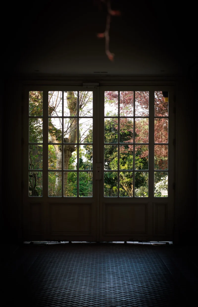 Claude · 3671 x 5685 Cette photographie montre une fenêtre à petits carreaux, sujet principal, encadrée par un mur sombre. La composition est épurée, avec un cadrage serré sur l'ouverture, laissant entrevoir un paysage arboré à l'extérieur.La lumière, tamisée et naturelle, filtre à travers les vitres, créant des contrastes entre ombres et clarté. Les couleurs dominantes sont les tons verts extérieurs et les noirs intérieurs, renforçant une ambiance mystérieuse et calme.Le style est minimaliste, évoquant une atmosphère contemplative et presque mélancolique.