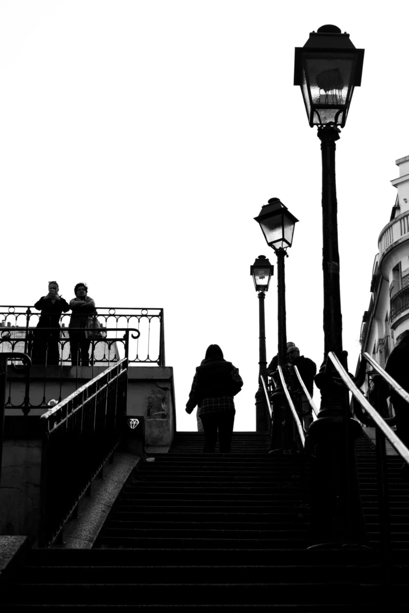 Claude · 3887 x 5830 Cette photographie en noir et blanc capture des silhouettes sur un escalier métallique urbain. Le sujet principal est un groupe de personnes descendant ou montant les marches, avec une personne assise en bas à droite.La composition repose sur des lignes verticales (lampadaires) et horizontales (escalier, garde-corps), créant une structure géométrique. Le cadrage est serré, mettant l’accent sur les contrastes d’ombres et de lumières.La lumière, douce et diffuse, accentue les contours et ajoute une ambiance mélancolique. Les tons dominants sont les noirs profonds et les gris, renforçant le côté dramatique et intemporel. Le style est épuré et cinématographique.