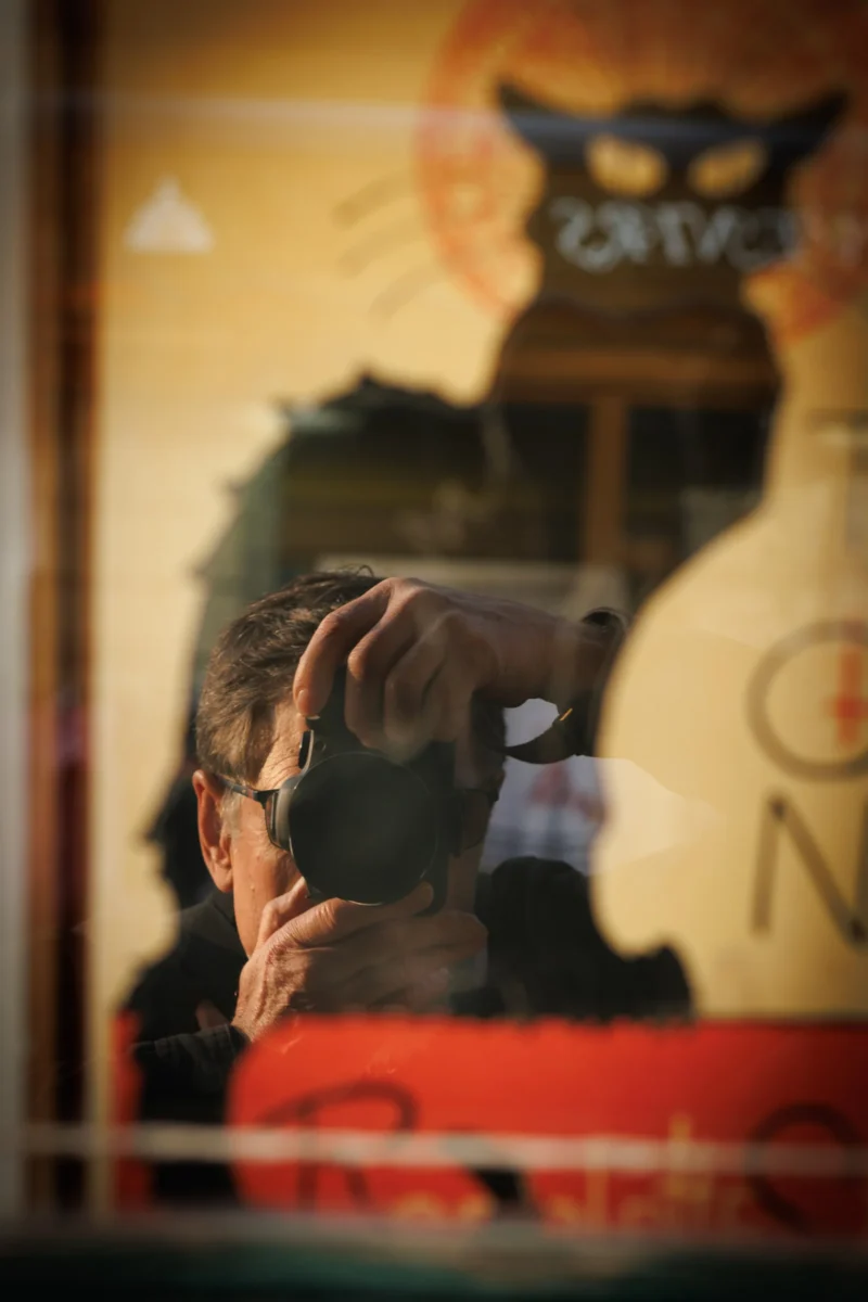 Claude · 2460 x 3690 Cette photographie met en scène un sujet principal flou, partiellement visible, avec une main couvrant son visage. Le cadrage est serré et centré sur cette silhouette, avec un arrière-plan légèrement déformé par une surface réfléchissante, comme une vitrine.La lumière est douce et tamisée, créant une ambiance intimiste et légèrement mélancolique. Les couleurs dominantes sont des tons chauds et sombres, avec des teintes orangées et brunes, accentuées par des reflets rouges.Le style est réaliste et naturel, évoquant une scène du quotidien. L'ambiance est à la fois mystérieuse et introspective.