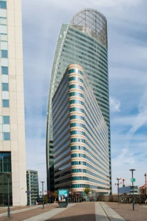Claude · 2495 x 3744 Cette photographie met en scène un **gratte-ciel moderne** aux formes courbes et à la façade vitrée, sujet principal de l'image. La **composition** joue sur la verticalité, accentuée par le cadrage serré et la perspective ascendante. Les **lignes architecturales** sont nettes, avec des jeux de transparence et de réflexion des vitres.La **lumière** est naturelle, douce et légèrement diffuse, créant des contrastes subtils. Les **couleurs dominantes** sont les tons gris bleutés des vitres, rehaussés par des touches de bleu et de blanc. Le style est **contemporain et urbain**, évoquant une ambiance **dynamique et sophistiquée**, typique d’un quartier d’affaires moderne.