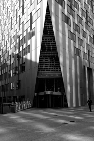 Claude · 3863 x 5794 Cette photographie met en scène un bâtiment moderne avec une entrée triangulaire en verre et métal. Le sujet principal est cette structure géométrique centrale, encadrée par deux tours résidentielles symétriques.La composition est épurée, axée sur les lignes droites et la symétrie. Le cadrage est serré, centré sur l’entrée.La lumière, naturelle et contrastée, crée des ombres marquées, accentuant les volumes. En noir et blanc, les tons dominants varient du blanc au gris foncé.Le style architectural est contemporain, minimaliste. L’ambiance est à la fois froide et élégante, évoquant une modernité urbaine et une certaine austérité.
