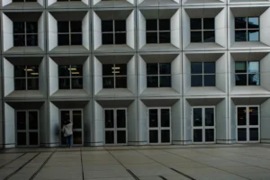 Claude · 5412 x 3608 Cette photographie met en scène un bâtiment moderne aux façades géométriques et répétitives, avec des fenêtres rectangulaires alignées. Le sujet principal est une personne en silhouette, marchant devant l'immeuble.La composition repose sur des lignes droites et une symétrie rigoureuse. Le cadrage est serré, centré sur la façade et la personne.La lumière est naturelle, tamisée, créant des contrastes doux. Les couleurs dominantes sont les gris et les tons neutres, renforçant une ambiance sobre et minimaliste. Le style est épuré, architectural, évoquant une froideur moderne et une atmosphère contemplative.