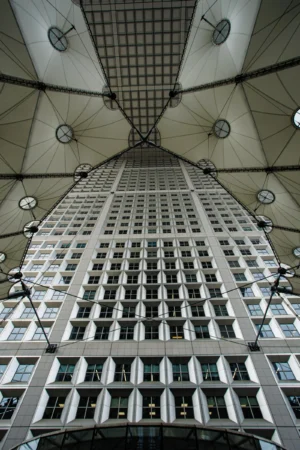 Claude · 3834 x 5751 Cette photographie met en scène un **immeuble moderne** vu depuis une **passerelle couverte**. Le **sujet principal** est l’architecture verticale, avec ses lignes épurées et ses fenêtres alignées.La **composition** joue sur la **perspective convergente**, créant un effet de profondeur et de symétrie. Le **cadrage** est serré, guidant le regard vers le haut.La **lumière** est naturelle, filtrée par la structure vitrée, apportant une ambiance claire mais contrastée. Les **couleurs dominantes** sont les tons gris-beige et blancs, renforçant le style **minimaliste et contemporain**.L’**ambiance** est à la fois **austère et futuriste**, évoquant une ville dynamique et architecturée.