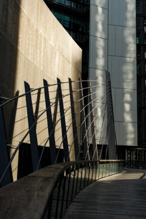 Claude · 3393 x 5090 Cette photographie met en scène un **escalier métallique moderne** avec des barrières en fil d’acier, intégré entre deux bâtiments contemporains. La **composition** joue sur les lignes droites et les jeux d’ombres. Le **cadrage** est serré, centré sur l’escalier et les reflets.La **lumière**, naturelle et rasante, crée des contrastes nets et des ombres marquées. Les **couleurs dominantes** sont des tons froids : gris, bleu et noir, avec des éclats de lumière dorée.Le style est **minimaliste et architectural**, évoquant une ambiance **urbaine et épurée**, presque industrielle.
