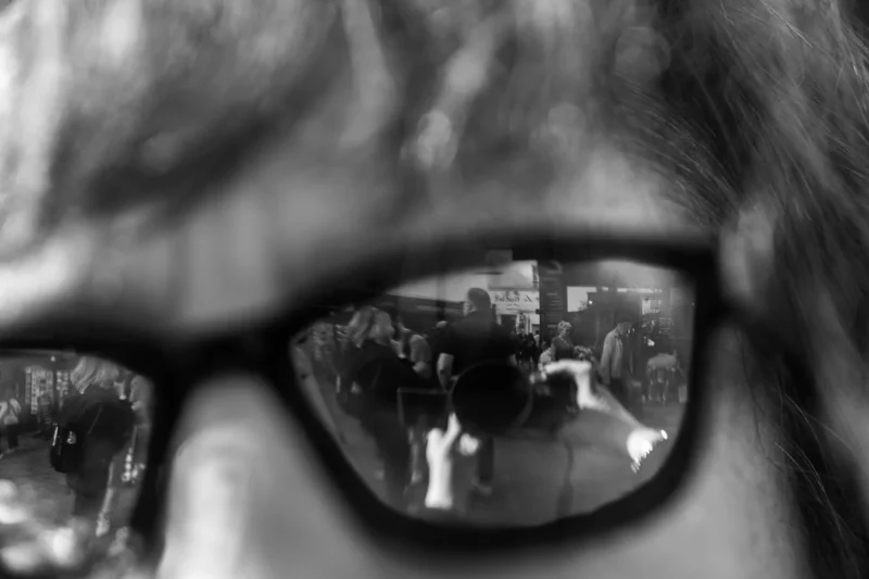 Laurence · 21/03/2026 · 1350 x 900 Cette photographie met en scène un **sujet principal flou**, vu à travers des **verres de lunettes**. La **composition** repose sur le reflet d’une rue animée, avec des silhouettes et des bâtiments.Le **cadrage** est serré, centré sur les lunettes, créant un effet de miroir intrigant. La **lumière** est douce, avec des contrastes marqués entre ombres et clartés. Les **couleurs dominantes** sont en noir et blanc, renforçant les textures et les jeux de lumière.Le **style** est minimaliste et artistique, évoquant une **ambiance introspective et urbaine**, mêlant intimité et anonymat.