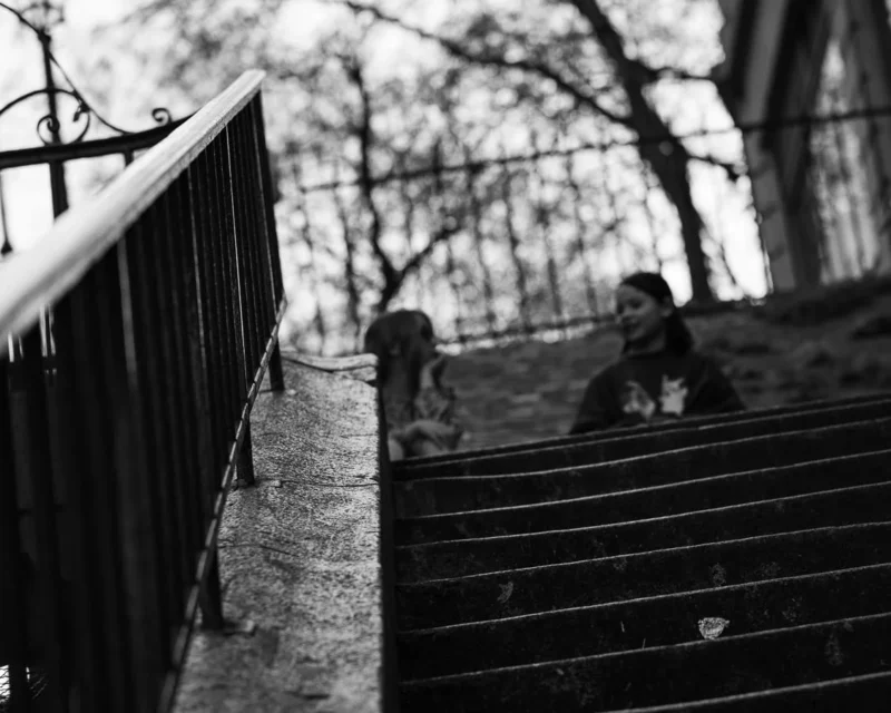 Laurence · 21/03/2026 · 1125 x 900 Cette photographie en noir et blanc met en scène deux personnes assises sur des marches extérieures, partiellement cachées par une rambarde. Le sujet principal est leur silhouette, évoquant une ambiance contemplative ou mélancolique.La composition joue sur les lignes verticales de la rambarde et horizontales des marches, créant une perspective profonde. Le cadrage serré et légèrement bas accentue ce sentiment d’intimité et d’isolement.La lumière douce et diffuse, probablement naturelle, adoucit les contours. Les tons gris dominants renforcent l’aspect nostalgique et intemporel. Le style est sobre et poétique, évoquant une ambiance calme et réfléchie.