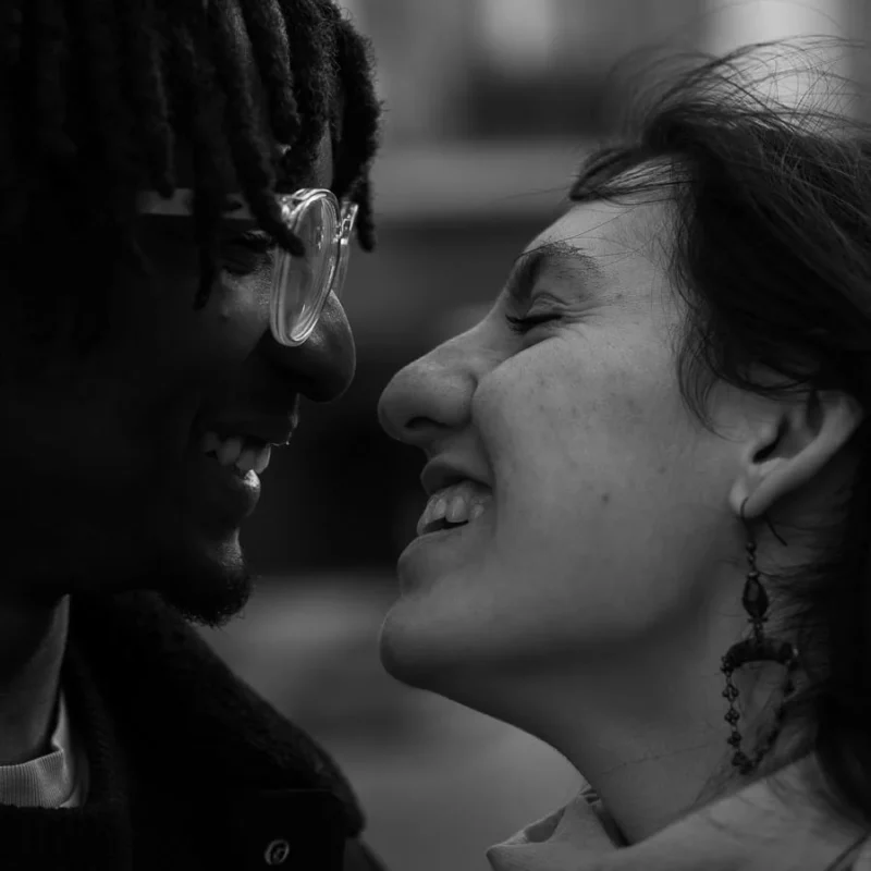 Laurence · 21/03/2026 · 900 x 900 Cette photographie met en scène deux personnes en train de rire, l’une masquée, l’autre souriante avec des boucles d’oreilles. Le sujet principal est leur complicité et leur joie partagée.La composition est serrée, centrée sur leurs visages, avec un cadrage en plan serré. La lumière est douce et naturelle, créant des contrastes doux sur leurs traits. En noir et blanc, les tons varient du clair au foncé, accentuant les expressions et les textures.Le style est documentaire et spontané, évoquant une ambiance chaleureuse et intime.