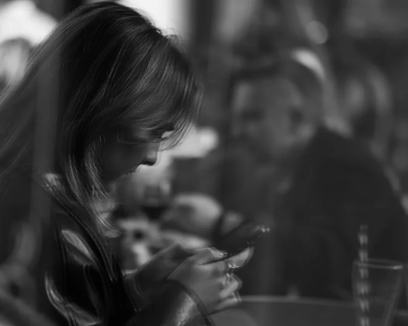 Laurence · 21/03/2026 · 1125 x 900 Cette photographie en noir et blanc met en scène une femme comme sujet principal, penchée sur son téléphone. La composition montre des personnes en arrière-plan, légèrement floues, créant une ambiance intimiste. Le cadrage serré isole la femme, attirant l’attention sur son expression concentrée.La lumière douce et tamisée accentue les contrastes, ajoutant une touche dramatique. Les couleurs dominantes, bien que limitées au noir et blanc, jouent sur les ombres et les clartés pour renforcer l’émotion.Le style est sobre et moderne, évoquant une ambiance contemplative et introspective.