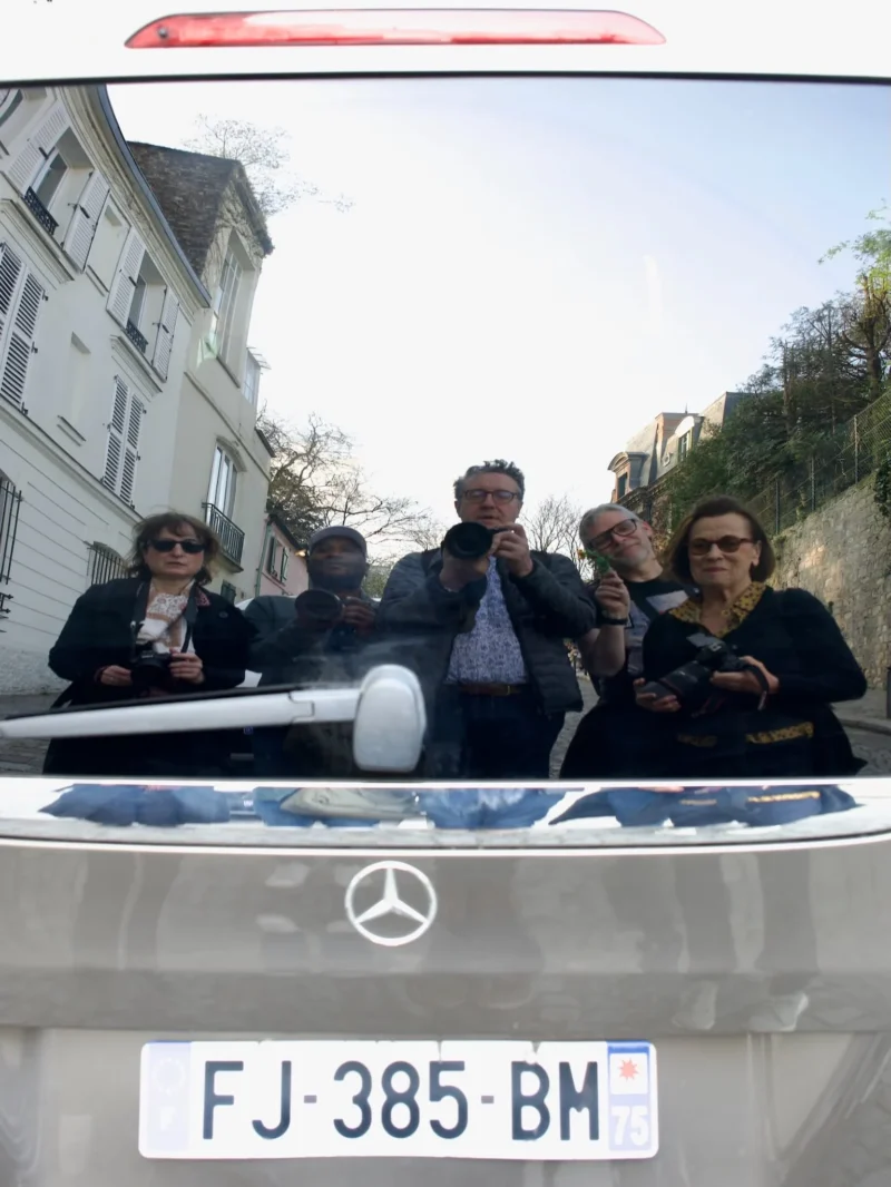 Xavier · 21/03/2026 · 1537 x 2048 Cette photographie montre un groupe de cinq personnes assises à l’arrière d’une Mercedes-Benz, visible dans le rétroviseur. Le sujet principal est le reflet des passagers, créant une composition symétrique et originale.Le cadrage est serré sur le pare-brise et la plaque d’immatriculation (FJ-385-BM). La lumière est naturelle, avec des ombres douces, suggérant un jour ensoleillé. Les couleurs dominantes sont les tons froids (bleu du ciel, gris de la voiture) et les teintes neutres des vêtements.Le style est documentaire et spontané, évoquant une ambiance décontractée et joyeuse.