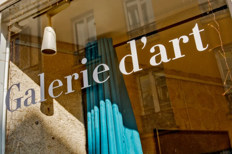 Frédéric · 21/03/2026 · 2517 x 1680 Cette photographie montre une vitrine d’une **galerie d’art**. Le sujet principal est l’inscription "Galerie d’art" en lettres blanches sur la vitre. La composition met en valeur cette enseigne, reflétée partiellement par la surface vitrée.Le cadrage est serré, centré sur la vitrine et le texte. La lumière naturelle éclaire la scène, créant des contrastes doux. Les couleurs dominantes sont les tons neutres (beige, gris) et les bleus froids du fond.Le style est épuré et moderne, évoquant une ambiance artistique et élégante. L’ensemble dégage une impression de raffinement et d’invitation culturelle.