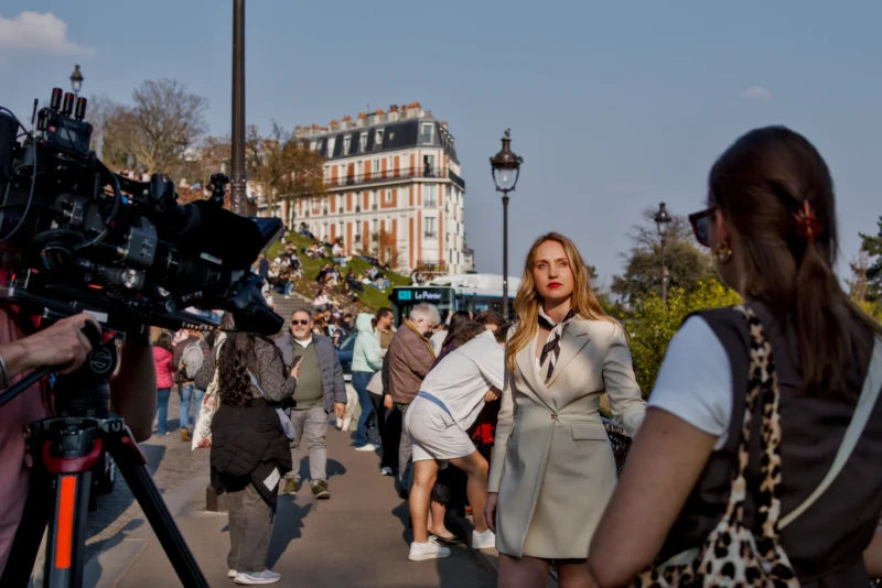 Frédéric · 21/03/2026 · 2517 x 1680 Cette photographie montre une femme en costume gris comme sujet principal, discutant avec une autre personne. La composition inclut plusieurs individus en arrière-plan, suggérant une scène publique ou un événement médiatique.Le cadrage est serré sur les deux femmes, avec une caméra de télévision visible à gauche. La lumière est naturelle et claire, typique d’un jour ensoleillé. Les couleurs dominantes sont des tons neutres (gris, beige) avec des touches de vert et de bleu dans l’arrière-plan.L’ambiance semble professionnelle et dynamique, évoquant une interview ou une couverture médiatique en extérieur.