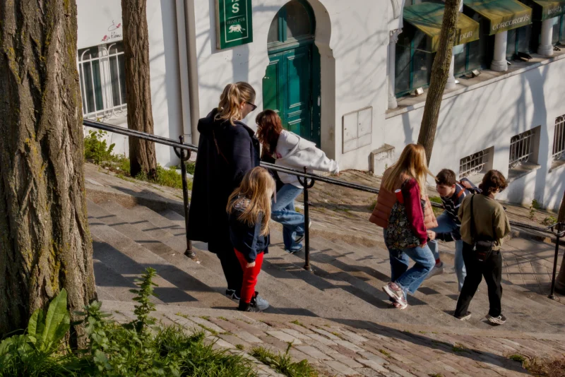 Frédéric · 21/03/2026 · 2517 x 1680 Cette photographie capture un groupe de personnes, principalement des enfants et des adultes, marchant sur une allée en pente pavée. Le sujet principal semble être les interactions entre ces individus.La composition est dynamique, avec un cadrage légèrement bas, mettant en valeur les visages et les mouvements. La lumière naturelle, douce et légèrement dorée, crée des ombres nettes. Les couleurs dominantes sont les tons neutres (beige, gris) avec des éclats de rouge et vert.Le style est documentaire et spontané, évoquant une ambiance chaleureuse et vivante dans un cadre urbain ou semi-urbain.