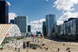 Yvon · 28/03/2026 · 6497 x 4332 Cette photographie montre une place urbaine moderne entourée de gratte-ciels. Le sujet principal est l’architecture contemporaine avec des bâtiments aux lignes épurées et des couleurs neutres. La composition met en valeur une large esplanade pavée, bordée de structures colorées et de drapeaux festifs.Le cadrage est large, capturant une vue panoramique. La lumière est naturelle, vive et éclatante, typique d’un jour ensoleillé. Les couleurs dominantes sont les gris des bâtiments, contrastés par les teintes vives des installations artistiques et des drapeaux. L’ambiance est dynamique et moderne, évoquant une ville en plein essor.