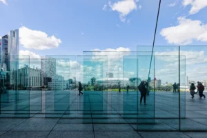 Yvon · 28/03/2026 · 6655 x 4437 Cette photographie met en scène une structure architecturale moderne en verre et métal, formant un cadre transparent. Le sujet principal est cette installation, reflétant les bâtiments environnants et le ciel.La composition joue sur les lignes droites et la symétrie, avec un cadrage serré qui isole cette structure. La lumière naturelle, vive et claire, accentue les reflets et les contrastes. Les couleurs dominantes sont les teintes bleutées du verre et les tons gris des bâtiments.Le style est épuré, minimaliste, typique de l’architecture contemporaine. L’ambiance est à la fois futuriste et apaisante, évoquant un espace urbain dynamique et réfléchissant.