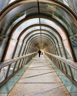 Yvon · 28/03/2026 · 3649 x 4561 Cette photographie montre un **passage couvert** avec une série d’**arcades métalliques** et une allée centrale en béton. Le **sujet principal** est une personne marchant au loin, créant une perspective profonde.La **composition** repose sur des lignes convergentes, renforçant l’effet de tunnel. Le **cadrage** est serré, mettant en valeur l’architecture et la symétrie. La **lumière** naturelle, tamisée, donne une ambiance douce et mystérieuse. Les **couleurs dominantes** sont des tons froids (bleutés, gris) avec des reflets verts.Le **style** est épuré et architectural, évoquant une **ambiance industrielle et contemplative**.