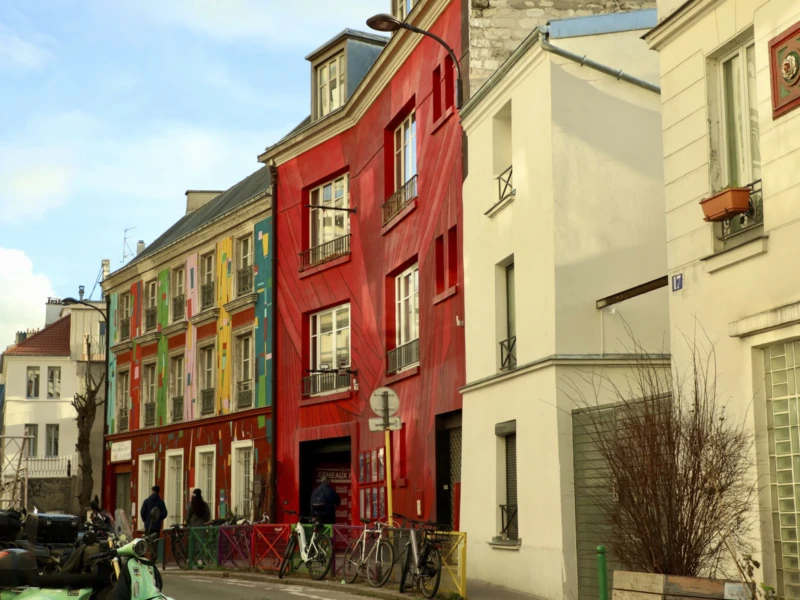 De Belleville à Ménilmontant Cette photographie montre une rue urbaine avec des bâtiments colorés comme sujet principal. La composition met en valeur une série d'immeubles aux façades variées, allant du rouge vif au jaune pâle et au blanc.Le cadrage est serré sur les maisons, avec une perspective légèrement inclinée. La lumière est naturelle, douce et diffuse, typique d’un jour légèrement nuageux. Les couleurs dominantes sont vives et contrastées, apportant une ambiance joyeuse et dynamique.Le style est réaliste et photographique, évoquant un quartier résidentiel pittoresque et animé. L’ambiance est chaleureuse et vivante, renforcée par les détails architecturaux et les touches de vie quotidienne.