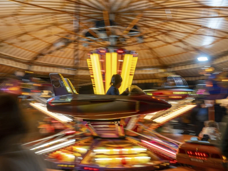 Flou Cette photographie capture un manège ou une attraction foraine en mouvement. Le sujet principal est le centre tourbillonnant des chevaux lumineux, floutés par la vitesse. La composition est dynamique, avec des lignes courbes et des couleurs vives. Le cadrage est serré, centré sur les chevaux illuminés.Les couleurs dominantes sont les tons chauds (jaunes, oranges) et les teintes pastel des chevaux. La lumière est artificielle et intense, créant une ambiance festive et légèrement onirique. Le style est photographique, avec un effet de mouvement suggéré par le flou. L’ambiance est joyeuse et énergique, typique d’une fête foraine.