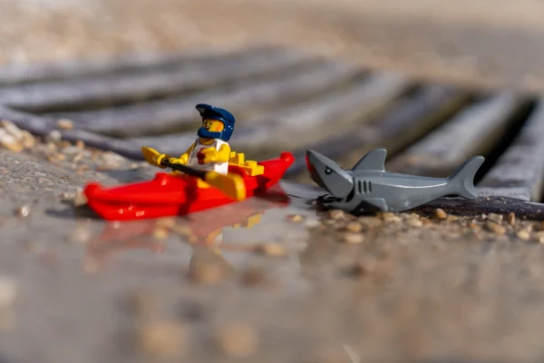 Marc Labbé · 14/03/2026 · 7008 x 4672 Cette photographie met en scène un petit personnage Lego vêtu d’un costume jaune et bleu, assis sur un objet rouge en forme de coque. À côté, un requin Lego gris semble émerger d’un fond flou.La composition est minimaliste, avec un cadrage serré sur les figurines. La lumière naturelle, douce et légèrement tamisée, crée des ombres douces. Les couleurs dominantes sont les tons vifs (jaune, bleu, rouge) contrastant avec le gris du requin et le fond neutre.Le style est épuré et ludique, évoquant une ambiance de jeu ou d’aventure miniature, avec une touche de mystère.