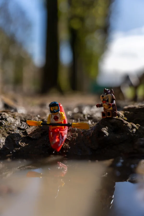 Marc Labbé · 14/03/2026 · 4672 x 7008 Cette photographie met en scène deux figurines de canoës-kayaks miniatures, dont l’une est rouge avec un personnage en combinaison bleue. Le sujet principal est le kayakiste en avant-plan, légèrement flou, tandis que l’autre kayakiste, en arrière-plan, est plus net.La composition joue sur la profondeur de champ, créant un effet de perspective. Le cadrage est serré, centré sur les figurines dans un ruisseau peu profond. La lumière naturelle, douce et légèrement tamisée, donne une ambiance calme et naturelle. Les couleurs dominantes sont les tons rouges, bleus et verts, renforçant une impression de nature et d’aventure. Le style est réaliste et documentaire, avec une touche artistique due au flou.