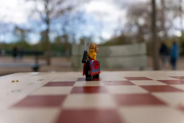 Marc Labbé · 14/03/2026 · 7008 x 4672 Cette photographie met en scène une figurine Lego habillée en costume, posée sur un damier rouge et blanc. Le sujet principal est net, tandis que l’arrière-plan est flou, créant un effet de profondeur.Le cadrage est serré, centré sur la figurine. La lumière naturelle semble douce, éclairant le sujet de face. Les couleurs dominantes sont les tons rouges et blancs du damier, avec des touches de vert et de gris en arrière-plan.Le style est épuré et minimaliste, évoquant une ambiance ludique et légèrement nostalgique.