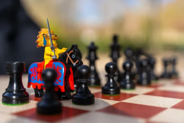 Marc Labbé · 14/03/2026 · 7008 x 4672 Cette photographie met en scène un **chessmen personnalisé** comme sujet principal : une pièce en forme de chevalier coloré (jaune et bleu) sur un cheval rouge, entourée de pièces noires classiques. La **composition** est serrée, centrée sur l’avant-plan du jeu d’échecs.Le **cadrage** est rapproché, avec un flou d’arrière-plan, mettant en valeur les détails. La **lumière** est naturelle et douce, créant des ombres légères. Les **couleurs dominantes** sont les noirs, rouges et blancs du plateau, contrastant avec les teintes vives de la pièce colorée.Le style est **créatif et ludique**, évoquant une ambiance **fantastique et joyeuse**.