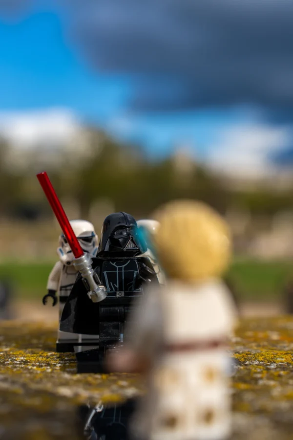 Marc Labbé · 14/03/2026 · 4672 x 7008 Cette photographie met en scène trois figurines LEGO représentant des personnages inspirés de *Star Wars*. Le sujet principal semble être les deux personnages en premier plan, l’un en tenue blanche (style Jedi) et l’autre en noir (style Sith), avec une épée rouge. Le cadrage est serré, mettant l’accent sur les détails des figurines. La lumière est naturelle, douce mais contrastée, créant des ombres marquées. Les couleurs dominantes sont les tons gris, blancs et noirs, avec des touches de rouge et bleu. Le style est minimaliste et ludique, évoquant une ambiance fantastique et épique.