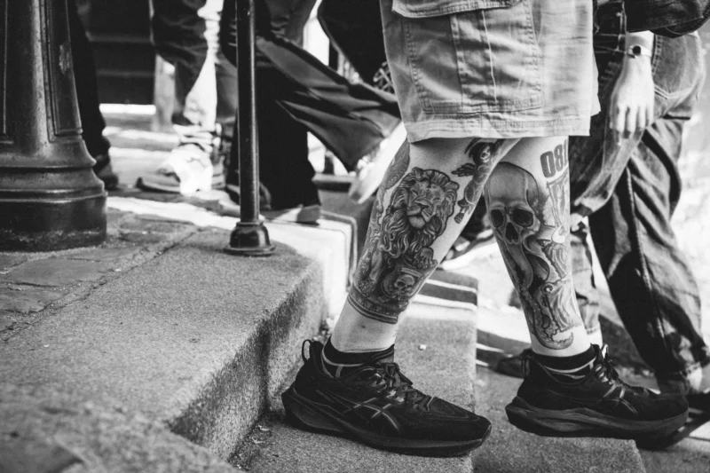 Marc Lab. · 21/03/2026 · 7008 x 4672 Cette photographie en noir et blanc met en valeur les **jambes et pieds d’une personne** assis·e sur une marche d’escalier. Le **sujet principal** est les tatouages détaillés sur les jambes, visibles grâce à un cadrage serré.La **composition** est simple et centrée, avec un jeu de lignes verticales (jambes) et horizontales (marche). La **lumière** naturelle, contrastée, accentue les textures et les motifs. L’**ambiance** est urbaine et intime, évoquant une scène de rue.Le style est **documentaire et artistique**, avec une touche de réalisme social. Les **tons de gris** dominent, renforçant l’aspect brut et authentique.