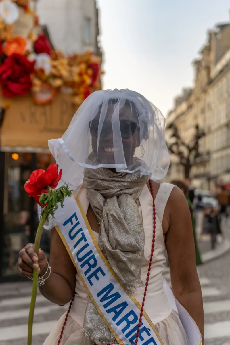 Marc Lab. · 21/03/2026 · 4261 x 6392 Cette photographie montre une personne vêtue d’une robe blanche et d’un voile, tenant une rose rouge. Son visage est masqué par un bandage, avec une inscription "FUTURE MARIÉE" sur un ruban bleu. Le cadrage est serré sur le sujet, avec un arrière-plan flou de bâtiments et de décorations festives.La lumière est naturelle, douce, mettant en valeur les couleurs pastel et le rouge vif de la rose. L’ambiance est à la fois mystérieuse et joyeuse, évoquant une célébration ou un événement festif. Le style est documentaire et artistique, avec une touche de théâtralité.