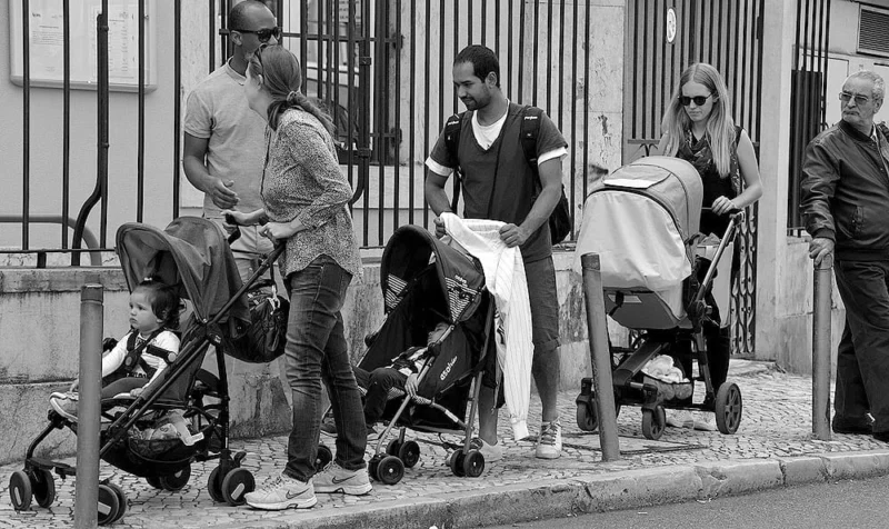 Pascale · 11/06/2018 · 1000 x 595 Cette photographie en noir et blanc capture un groupe de personnes en ville. Le sujet principal semble être une famille avec des enfants en bas âge, dont les poussettes sont au premier plan.La composition montre un cadrage serré sur le trottoir, avec une mise en scène dynamique et naturelle. La lumière est douce, probablement naturelle, créant des contrastes modérés.L’ambiance est quotidienne et réaliste, reflétant une scène urbaine paisible. Le style est documentaire, mettant l’accent sur la vie de tous les jours. Les tons de gris dominent, renforçant l’aspect authentique et intemporel de la scène.