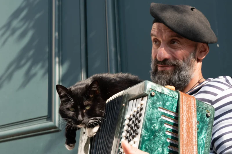 Alice · 21/03/2026 · 3896 x 2597 Cette photographie met en scène un homme barbu portant un béret et un pull coloré, tenant un chat noir dans ses bras. Le sujet principal est l’homme et son interaction avec l’animal, créant une ambiance chaleureuse et naturelle.La composition est simple et centrée, avec un cadrage serré qui isole les deux sujets. La lumière semble naturelle, douce et légèrement latérale, adoucissant les traits du visage et du chat. Les couleurs dominantes sont les tons verts et orange du pull, contrastant avec le noir du chat et le gris du fond. Le style est réaliste et spontané, évoquant une scène du quotidien.