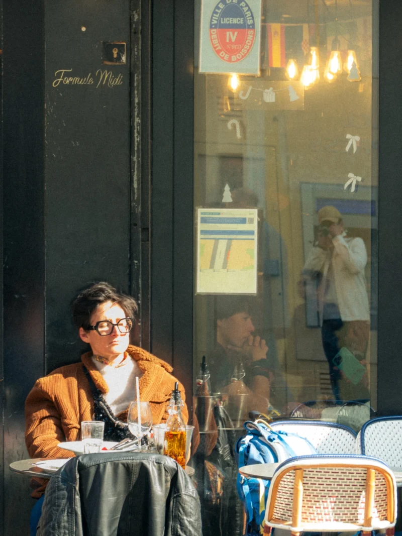 Seila · 21/03/2026 · 2592 x 3456 Cette photographie montre une personne assise à une table à l’intérieur d’un café, vêtue d’un manteau orange et de lunettes. Le sujet principal est cette personne, en contre-jour partiel, reflétée dans la vitre.La composition joue sur la transparence de la vitre, intégrant des éléments extérieurs et intérieurs. Le cadrage est serré, mettant en valeur les détails du visage et de l’environnement immédiat.La lumière est douce et naturelle, avec des tons chauds. Les couleurs dominantes sont les teintes neutres et l’orange du manteau. L’ambiance est calme et introspective, évoquant une pause contemplative dans un cadre urbain.