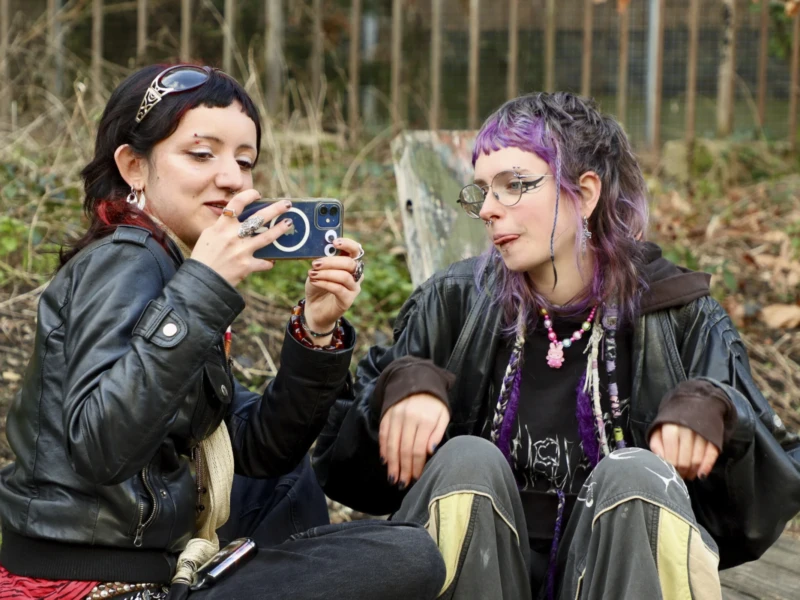 Portraits de rue Cette photographie met en scène deux jeunes femmes assises en extérieur. Le sujet principal est leur interaction : l’une montre quelque chose à l’autre avec enthousiasme, probablement un appareil photo.La composition est naturelle et spontanée, avec un cadrage serré sur les visages et les épaules. La lumière semble douce et naturelle, typique d’un jour nuageux. Les couleurs dominantes sont des tons neutres (verts de la nature, gris et marron) avec des touches de noir (vêtements) et de violet (accessoires).Le style est décontracté et authentique, évoquant une ambiance amicale et détendue.