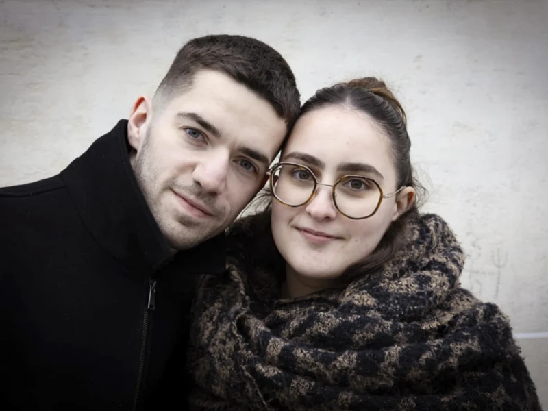 Portraits de rue Cette photographie met en scène un couple en plan serré, sujet principal. Le cadrage est rapproché, centré sur leurs visages et épaules. La composition est symétrique et intime, avec un léger contact entre leurs têtes.La lumière est douce et naturelle, créant des ombres subtiles. Les couleurs dominantes sont les tons neutres : gris, noir et beige, adoucis par les reflets des vêtements.Le style est épuré et réaliste, évoquant une ambiance chaleureuse et sincère, presque minimaliste. L'ensemble dégage une impression de complicité et de simplicité.