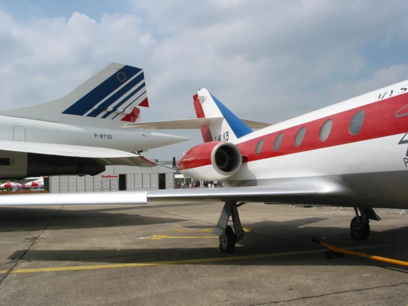 Frédéric · 15/06/2003 · 1600 x 1200 Cette photographie montre deux avions au sol sur un tarmac. Le sujet principal est un avion blanc et rouge avec des détails bleus, partiellement visible, tandis qu’un autre avion aux couleurs bleu-blanc-rouge (Air France) apparaît en arrière-plan.La composition met en valeur les avions avec un cadrage serré sur leurs extrémités. La lumière est naturelle, légèrement diffuse, créant des contrastes doux. Les couleurs dominantes sont le blanc, le rouge et le bleu, renforçant l’identité des compagnies aériennes. Le style est documentaire, captant une scène aéroportuaire réaliste. L’ambiance est professionnelle et industrielle, typique des aéroports.