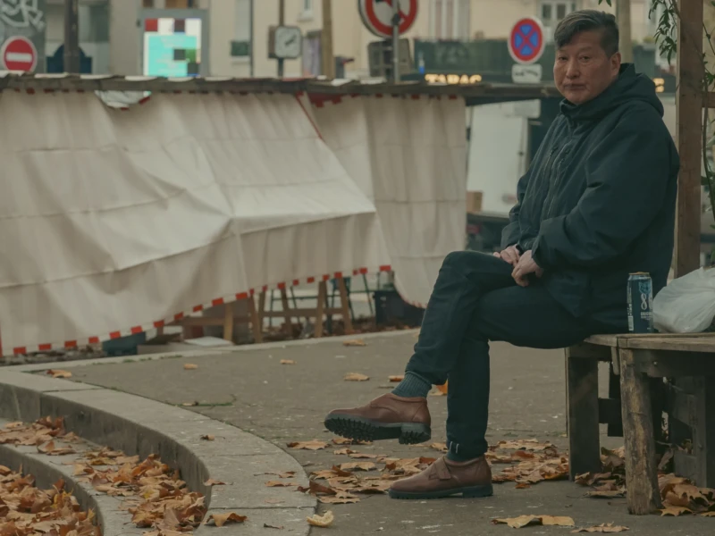 Village de Charonne Cette photographie montre un homme assis seul sur un banc, dans une posture détendue mais légèrement absorbée. Le sujet principal est mis en valeur par un cadrage serré, centré sur lui.La composition inclut un fond urbain avec des éléments comme des tentes et des feuilles mortes. La lumière semble naturelle, tamisée, créant des ombres douces. Les couleurs dominantes sont des tons froids (bleu du manteau, gris de l’asphalte) et des touches de terre (feuilles, sol).Le style est réaliste et sobre, évoquant une ambiance calme et mélancolique, presque introspective.