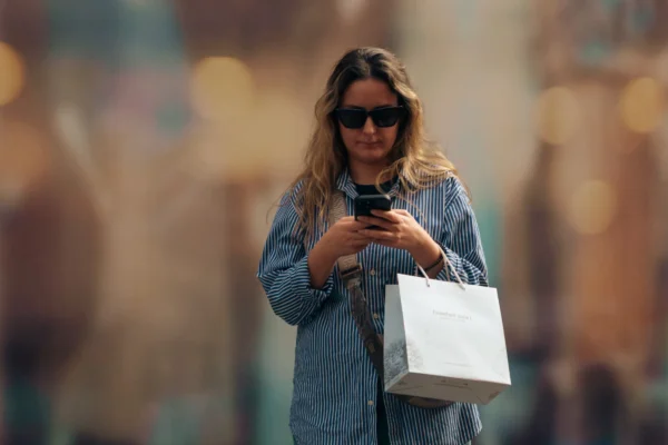 Don SOUCEMARIANADIN · 07/03/2026 · 2984 x 1990 Cette photographie met en scène une femme comme sujet principal, vêtue d'une veste bleu clair, tenant un téléphone et un sac blanc. La composition est simple, avec un cadrage serré sur elle, légèrement flou en arrière-plan.La lumière est douce et naturelle, créant des reflets et des bokehs colorés. Les couleurs dominantes sont les tons bleus et blancs, avec des éclats chauds en arrière-plan. Le style est contemporain et urbain, évoquant une ambiance moderne et dynamique, presque intimiste malgré la présence de gens en arrière-plan.
