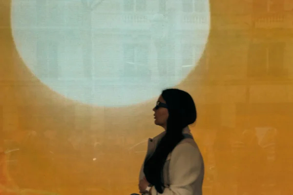 Don SOUCEMARIANADIN · 07/03/2026 · 4646 x 3097 Cette photographie met en scène une femme comme sujet principal, vue de profil, les yeux légèrement fermés ou perdus dans le lointain. La composition est épurée, avec un cadrage serré sur elle, devant une grande surface lumineuse en forme de cercle.La lumière est douce et diffuse, créant une ambiance contemplative. Les couleurs dominantes sont des tons chauds (beige, orange) et une teinte froide bleutée pour le cercle lumineux. Le style est sobre et minimaliste, évoquant une sensation de calme et de réflexion. L’ambiance est introspective et presque onirique.