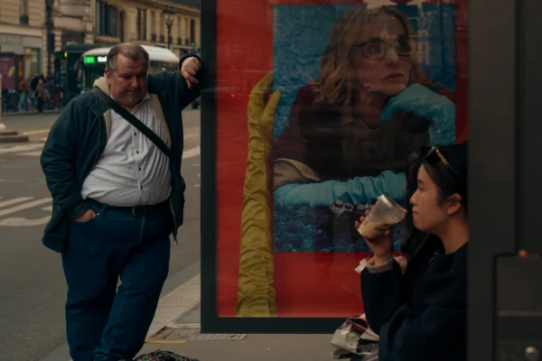 Don SOUCEMARIANADIN · 07/03/2026 · 4356 x 2904 Cette photographie montre un homme en surpoids, vêtu d’un pull blanc et d’un pantalon bleu, posant devant une affiche publicitaire rouge. Une femme, partiellement visible, semble manger un sandwich en bas à droite.La composition joue sur le contraste entre le sujet principal et l’affiche colorée. Le cadrage est serré, mettant en valeur les détails des visages et des vêtements. La lumière est naturelle, légèrement tamisée, créant des ombres douces. Les couleurs dominantes sont les tons rouges, bleus et gris, renforçant une ambiance urbaine et réaliste. Le style est documentaire, capturant un instant de vie quotidienne avec authenticité.
