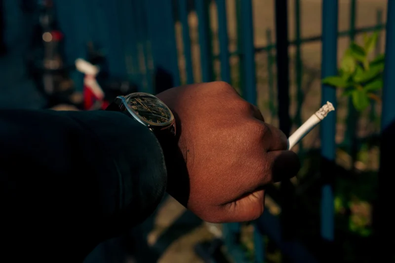 Don · 21/03/2026 · 2048 x 1366 Cette photographie met en scène une main tenant une cigarette allumée, avec une montre visible au poignet. Le sujet principal est donc cette main, partiellement vêtue d’un pull noir.Le cadrage est serré, centré sur la main et l’objet, avec un arrière-plan flou montrant une barrière et des éléments urbains. La lumière est tamisée, créant des contrastes doux et une ambiance nocturne ou peu éclairée. Les couleurs dominantes sont les teintes sombres (noir, gris) avec des touches de bleu et de vert en arrière-plan.Le style est réaliste et sobre, évoquant une ambiance contemplative et intime.