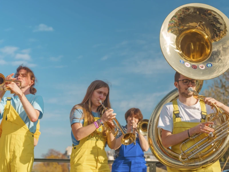 De Belleville à Ménilmontant Cette photographie met en scène un groupe de jeunes musiciens jouant des instruments à vent. Le sujet principal est un tuba, entouré de trompettes et d'un trombone, avec des musiciens vêtus de tenues jaunes.La composition est dynamique, avec un cadrage serré sur les instruments et les visages des musiciens. La lumière est naturelle, probablement en extérieur, avec un éclairage doux et homogène. Les couleurs dominantes sont le jaune vif des vêtements et les tons dorés des instruments, contrastant avec le bleu du ciel.Le style est documentaire et joyeux, évoquant une ambiance conviviale et festive.