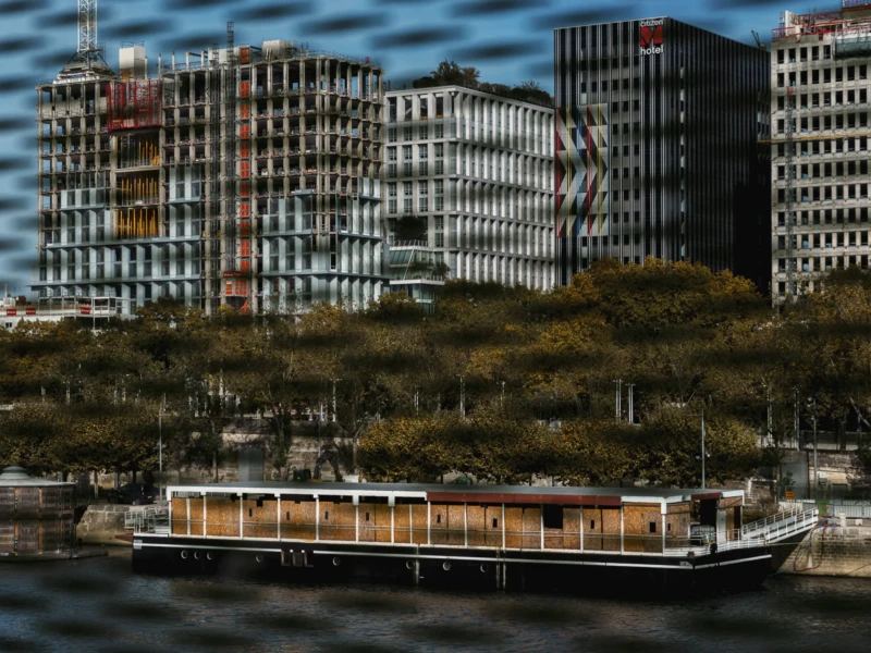 Paris et ses lieux oubliés Cette photographie met en scène un bateau à roues sur un plan d’eau, en premier plan. L’arrière-plan montre une ville avec des bâtiments modernes et une colline boisée.La composition joue sur les contrastes entre le bateau traditionnel et l’architecture contemporaine. Le cadrage est légèrement flou, avec une perspective qui donne une impression de profondeur.Les couleurs dominantes sont les tons gris et bruns des bâtiments, contrastant avec le vert des arbres et le bleu de l’eau. La lumière semble naturelle, avec des ombres douces, créant une ambiance à la fois urbaine et nostalgique.