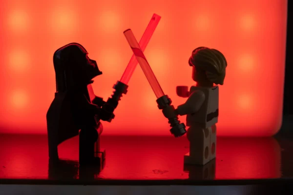 Juan MEDINA MARTINEZ · 7581 x 5057 Cette photographie met en scène deux figurines inspirées de personnages de *Star Wars* : Darth Vader et un jeune Jedi (probablement Luke Skywalker enfant). La composition montre un duel stylisé avec des sabres laser croisés.Le cadrage est serré, centré sur les personnages. La lumière, rougeâtre et diffuse, crée une ambiance dramatique et mystérieuse. Les couleurs dominantes sont le rouge et le noir, renforçant l'atmosphère sombre et intense. Le style évoque une scène cinématographique, avec un éclairage théâtral. L'ambiance est épique et chargée d'émotion.