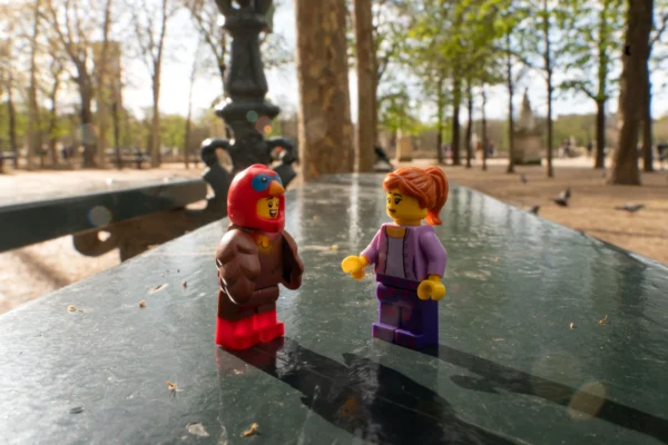 Juan MEDINA MARTINEZ · 6865 x 4579 Cette photographie met en scène deux figurines Lego comme sujets principaux. Elles sont placées sur une surface noire et humide, probablement du métal ou du verre.La composition est symétrique et minimaliste, avec un cadrage serré sur les figurines. L’arrière-plan montre un parc avec des arbres et des personnes floutées, créant une ambiance urbaine et détendue.La lumière est naturelle et douce, avec des ombres discrètes. Les couleurs dominantes sont les tons vifs des vêtements des figurines (rouge, violet) et les teintes vertes des arbres en arrière-plan. Le style est épuré et ludique, évoquant une touche de fantaisie dans un cadre réaliste.