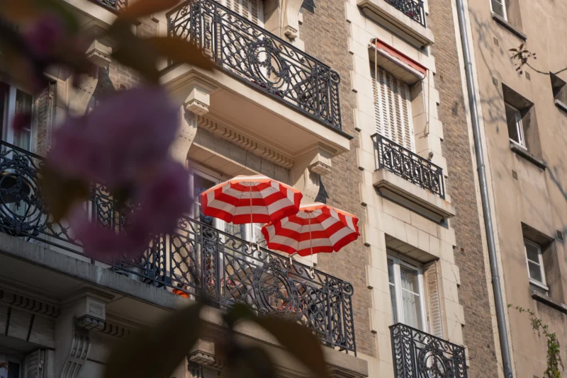 Juan · 21/03/2026 · 7280 x 4856 Cette photographie met en scène des **parasols rouges** accrochés à une façade d’immeuble parisien. Le **sujet principal** est ces parasols, disposés en série sur un balcon.La **composition** joue sur le contraste entre les éléments naturels (fleurs en premier plan) et urbains (bâtiment ancien). Le **cadrage** est serré, avec une légère flou artistique, mettant l’accent sur les couleurs vives.Les **couleurs dominantes** sont le rouge des parasols, le vert des feuilles et le beige de la pierre. La **lumière** est douce, naturelle, créant une ambiance **romantique et estivale**. Le style évoque un **Paris pittoresque et bohème**.