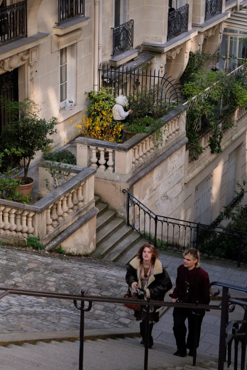Juan · 21/03/2026 · 4300 x 6447 Cette photographie montre deux personnes descendant un escalier en pierre dans un cadre urbain. Le sujet principal est les deux individus, l’un portant un sac à main et l’autre un sac à dos.La composition met en valeur les escaliers et les balcons fleuris d’un immeuble parisien typique. Le cadrage est légèrement incliné, offrant une perspective dynamique. La lumière est douce et naturelle, probablement en fin de journée, créant des ombres douces.Les couleurs dominantes sont les tons neutres (beige, gris) avec des touches de vert des plantes et de jaune des fleurs. Le style est documentaire et réaliste, évoquant une ambiance calme et quotidienne dans un quartier résidentiel.