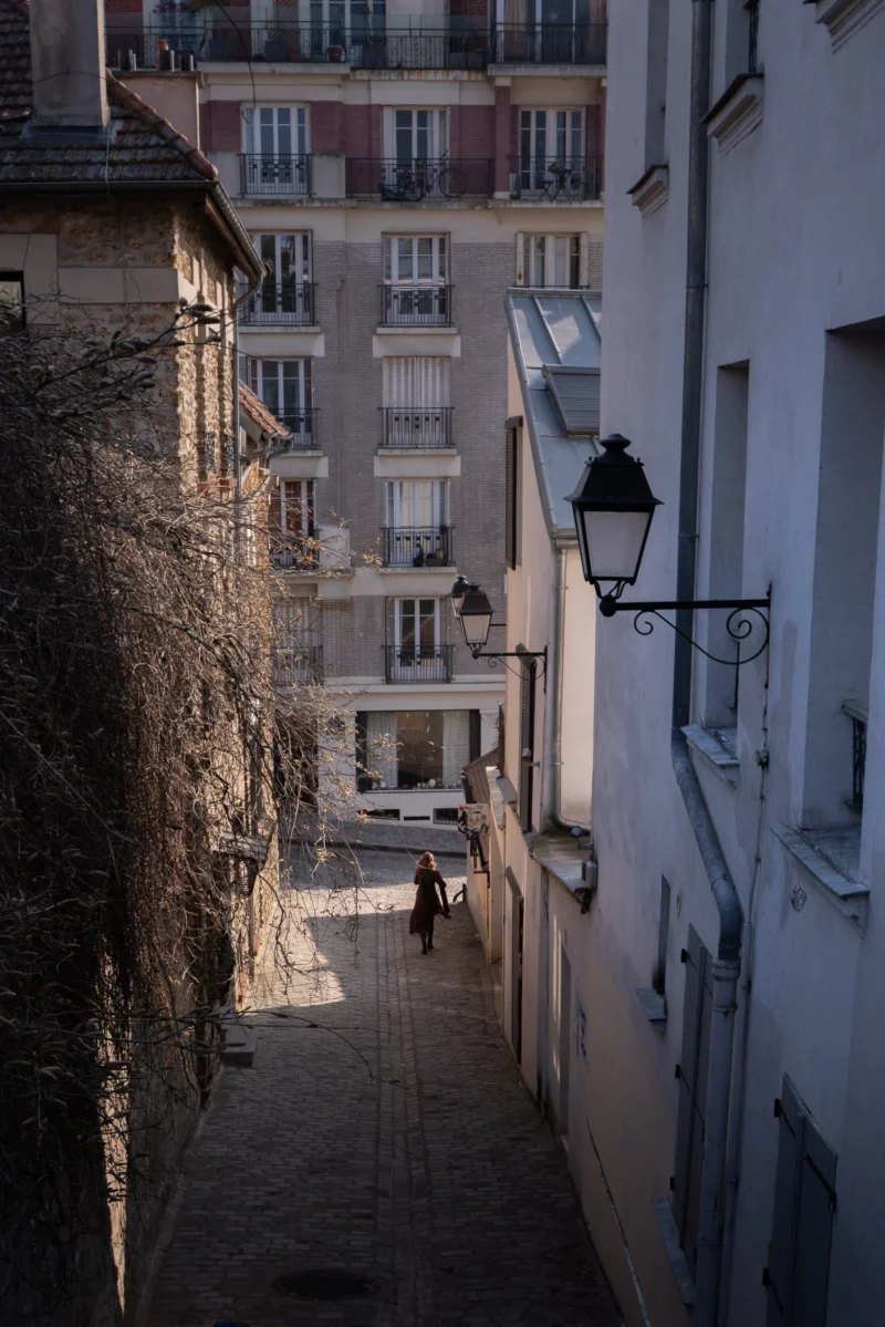 Juan · 21/03/2026 · 2990 x 4483 Cette photographie capture une ruelle étroite et sinueuse en ville. Le sujet principal est l’allée descendante bordée de bâtiments anciens, avec une personne en contrebas. La composition repose sur des lignes convergentes menant vers le fond, renforçant la perspective.Le cadrage est serré, mettant en valeur les détails architecturaux et l’ivraie grimpante. La lumière, douce et tamisée, crée des ombres longues et une ambiance calme et mélancolique. Les couleurs dominantes sont des tons neutres : gris, beige et blanc, adoucis par des touches de vert.Le style est documentaire et poétique, évoquant une atmosphère paisible et intemporelle.