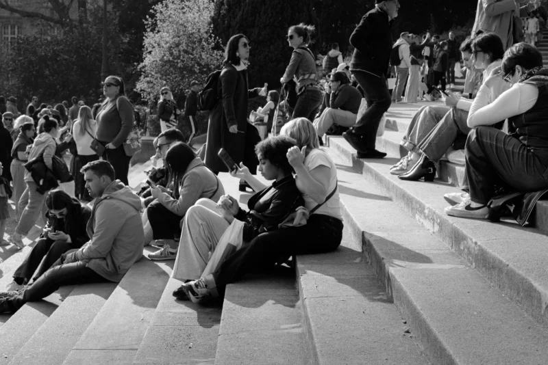 Juan · 21/03/2026 · 7776 x 5187 Cette photographie en noir et blanc capture un groupe de personnes assises sur des marches extérieures, semblant discuter ou se reposer. Le sujet principal est l'interaction sociale dans un espace public.La composition montre une disposition en gradins, avec des individus répartis sur plusieurs niveaux. Le cadrage est large, incluant un arrière-plan flou avec des arbres et des personnes debout. La lumière naturelle, probablement en journée, crée des contrastes nets et des ombres marquées. L'absence de couleurs renforce l'aspect intemporel et universel de la scène, évoquant une ambiance calme et détendue. Le style est documentaire et réaliste.